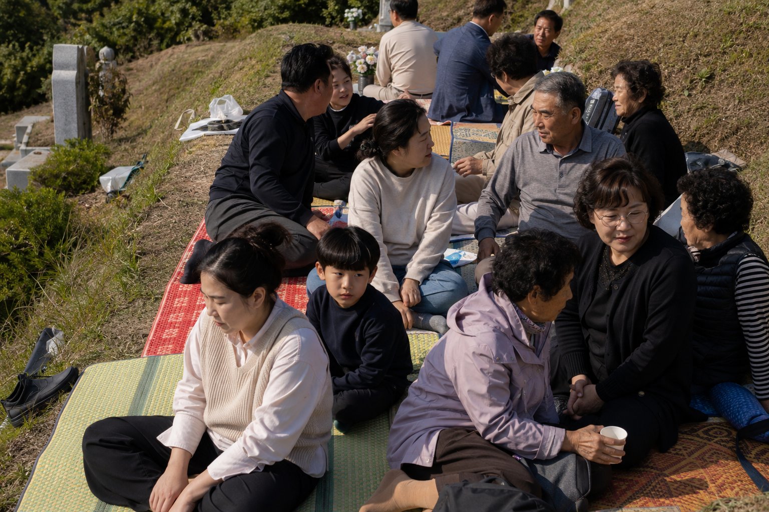 좁은 공동묘지에 여러 명이 앉아 있고 돗자리들이 겹쳐있는 모습